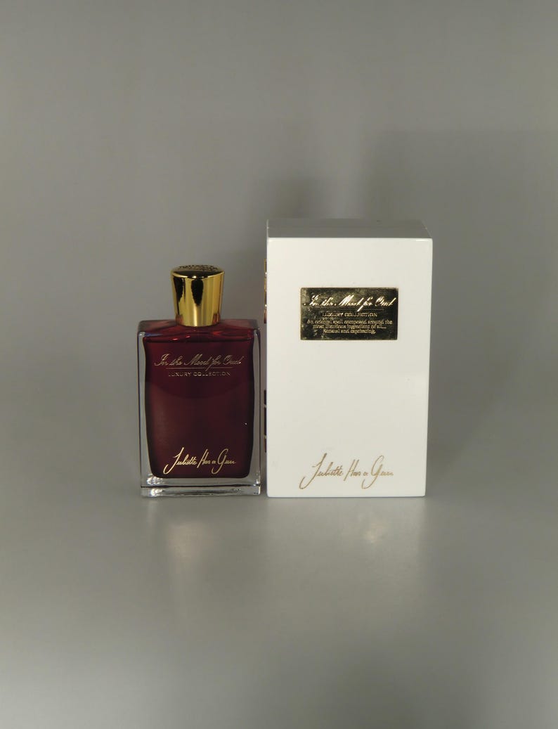 Op de afbeelding: Een glazen fles parfum met een gouden dop en een rode vloeistof. De fles is gelabeld met "La Plus Belle" en "Juliette Has a Gun". De fles staat op een wit oppervlak naast een witte doos met hetzelfde label.