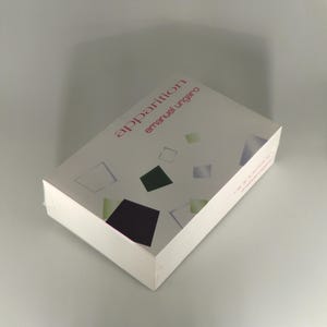 May include: A white book titled "apparition" by Emanuel Ungaro. The cover features geometric shapes in black, green, and blue. The book is wrapped in clear plastic.