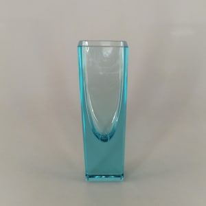 Puede incluir: Un jarrón de vidrio azul claro con una base cuadrada y una abertura redondeada.