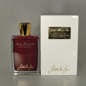 Op de afbeelding: Een doorzichtige glazen fles parfum met een gouden dop en een rode vloeistof. De fles is gelabeld met "In the Mood for Oud" en "Juliette Has a Gun" in gouden letters. De fles staat op een wit oppervlak naast een witte doos met hetzelfde label.