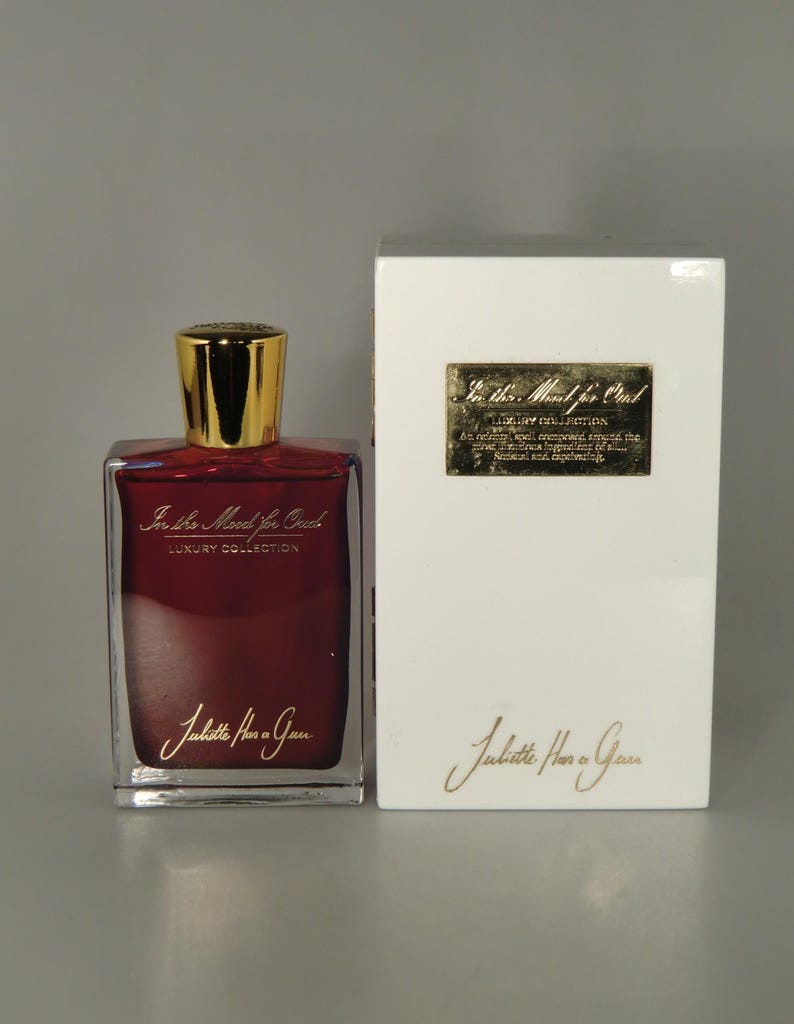 Op de afbeelding: Een doorzichtige glazen fles parfum met een gouden dop en een rode vloeistof. De fles is gelabeld met "In the Mood for Oud" en "Juliette Has a Gun" in gouden letters. De fles staat op een wit oppervlak naast een witte doos met hetzelfde label.