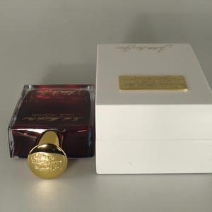 Op de afbeelding: Een donkerrode glazen fles parfum met een gouden dop en etiket. De fles staat op een witte doos met de tekst "The Alchemist" erop.