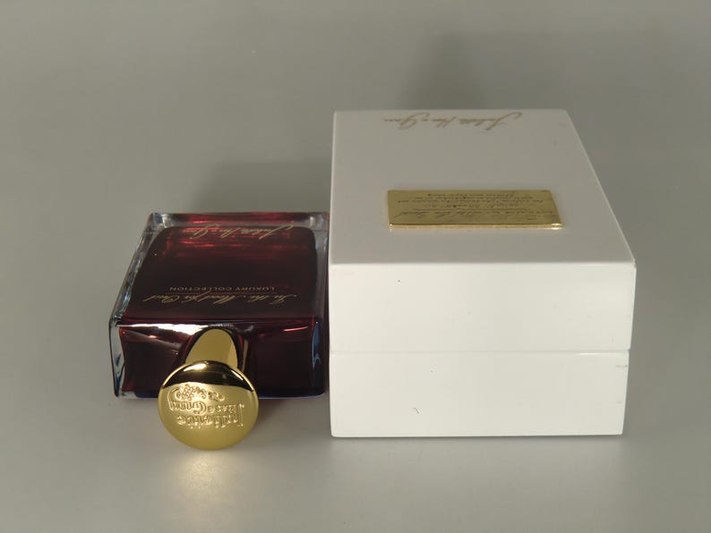 Op de afbeelding: Een donkerrode glazen fles parfum met een gouden dop en etiket. De fles staat op een witte doos met de tekst "The Alchemist" erop.