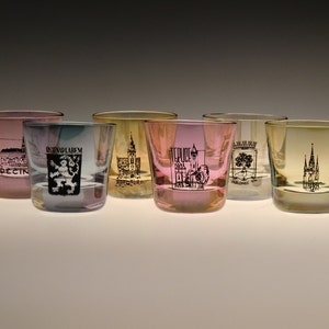 Puede incluir: Seis pequeños vasos de chupito de vidrio transparente con diferentes matices de color. Cada vaso tiene una imagen en blanco y negro de un edificio o escudo en la parte delantera. Los vasos están dispuestos en fila sobre una superficie blanca.