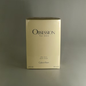 May include: A light beige box of Calvin Klein Obsession aftershave for men. The box features the product name and brand name in black. The box contains 125 ml of aftershave.