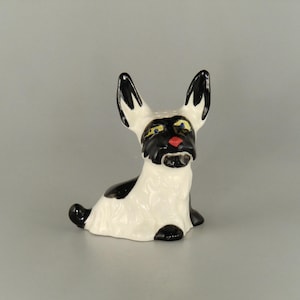 Puede incluir: Una figura de cerámica de un perro, principalmente blanca con detalles en negro. El perro tiene orejas grandes y erguidas con puntas negras, una cara negra con ojos amarillos y una nariz roja. El perro está sentado.