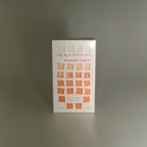 May include: A white box of Emanuel Ungaro's "apparition sun" eau de toilette. The box features the brand name and product name in a pink font. The word "sun" is displayed in orange squares. The box contains 90 ml and 3 fl oz.