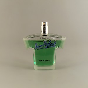 May include: A clear glass bottle of Sonia Rykiel L'Eau de Parfum. The bottle is shaped like a t-shirt and contains a green liquid. The label on the bottle reads "L'Eau de Sonia Rykiel" in blue lettering.