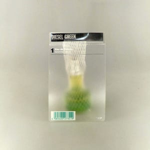 May include: A clear rectangular perfume box with the text "DIESEL GREEN" in black and white. The box contains a perfume bottle with a green base and a white net-like covering. A barcode is visible at the bottom.
