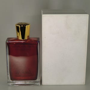 Op de afbeelding: Een heldere glazen fles met een gouden dop gevuld met een donkerrode vloeistof. De fles staat naast een witte doos.