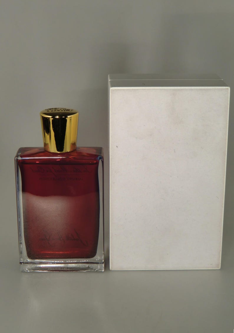 Op de afbeelding: Een heldere glazen fles met een gouden dop gevuld met een donkerrode vloeistof. De fles staat naast een witte doos.