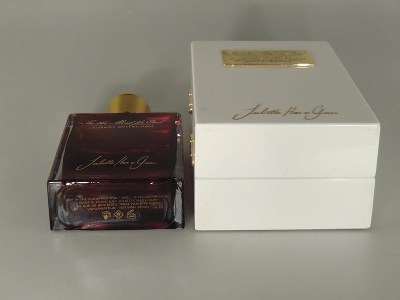 Op de afbeelding: Een donkerrode glazen fles parfum met een gouden dop en een witte doos. De fles heeft de tekst "Juliette Has a Gun" erop. De doos heeft de tekst "Juliette Has a Gun" erop.