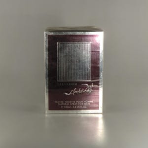 May include: A rectangular perfume box with a silver frame and a burgundy centre panel. The panel features a textured silver square and the name "SALVADOR" above the signature of Salvador Dalí. The text "EAU DE TOILETTE POUR HOMME NATURAL SPRAY FOR MEN e 100ml - 3.4 US FL.OZ." is printed below.