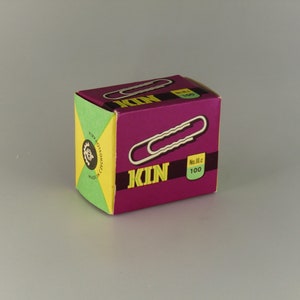 Könnte beinhalten: Eine Schachtel mit 100 silbernen Büroklammern mit dem Wort "KIN" in Gelb und Lila auf der Vorderseite. Die Schachtel ist grün, gelb und lila.