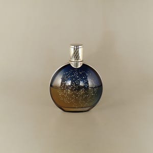 May include: A round perfume bottle with a silver cap. The glass bottle transitions from a dark blue at the top to a golden hue at the bottom. White constellations are printed on the glass.