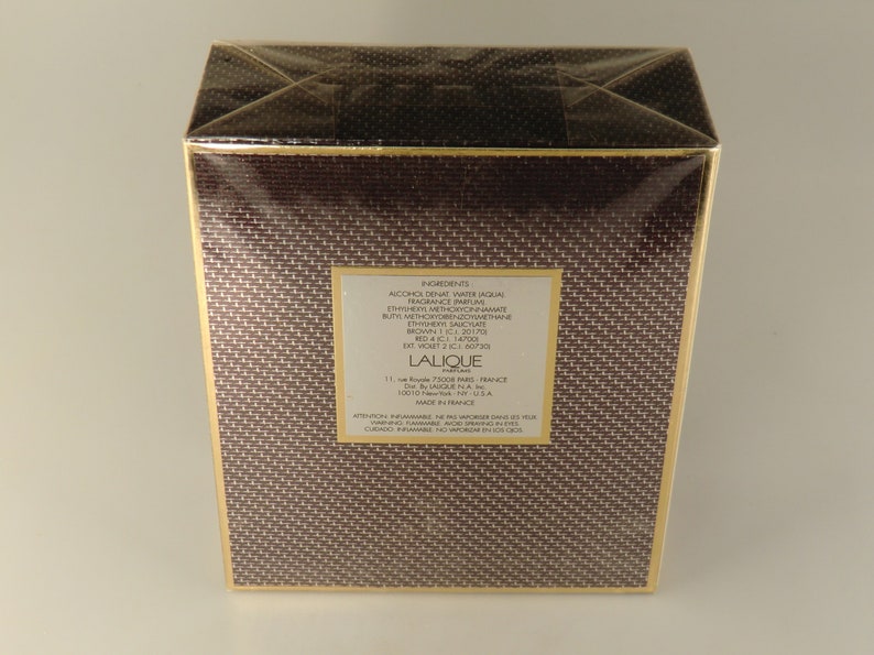 K&ouml;nnte beinhalten: Eine braune Schachtel mit goldenen Verzierungen und einem wei&szlig;en Etikett, auf dem die Inhaltsstoffe des Lalique-Parf&uuml;ms aufgef&uuml;hrt sind. Das Etikett enth&auml;lt den Text "Lalique Perfume" und "Made in France".
