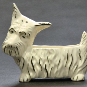 Może przedstawiać: Biała ceramiczna doniczka w kształcie szorstkowłosego teriera szkockiego. Doniczka ma czarne detale i długi, wąski otwór do sadzenia.