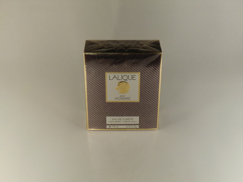 K&ouml;nnte beinhalten: Eine braune und goldene Schachtel mit dem Text "Lalique Pour Homme" und "Eau de Toilette Natural Spray" darauf. Die Schachtel ist 75 ml oder 2,5 fl oz.