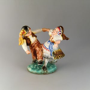Peut inclure: Une figurine en céramique représentant un garçon et une fille dansant en costumes folkloriques traditionnels. Le garçon porte un gilet et un pantalon bruns, tandis que la fille porte une robe blanche avec des accents bleus et rouges. La figurine est peinte dans des couleurs vives et a une base verte.
