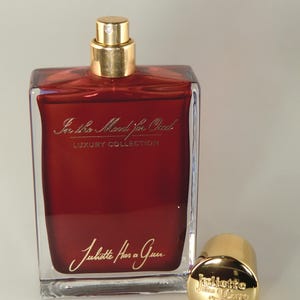 Op de afbeelding: Een doorzichtige glazen fles parfum met een gouden dop en een rode vloeistof erin. De fles is gelabeld met "In the Mood for Oud" en "Juliette Has a Gun" in gouden letters. De fles staat op een wit oppervlak.