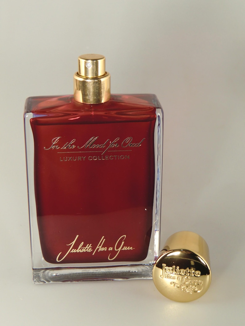 Op de afbeelding: Een doorzichtige glazen fles parfum met een gouden dop en een rode vloeistof erin. De fles is gelabeld met "In the Mood for Oud" en "Juliette Has a Gun" in gouden letters. De fles staat op een wit oppervlak.