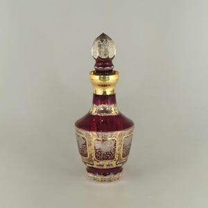 Könnte beinhalten: Eine dekorative, rubinrote Glasflasche mit goldfarbenem Rand und Verschluss. Die Flasche hat ein verziertes Design mit goldenen Akzenten und einen klaren, spitz zulaufenden Verschluss. Die Flasche ist etwa 15 cm hoch.