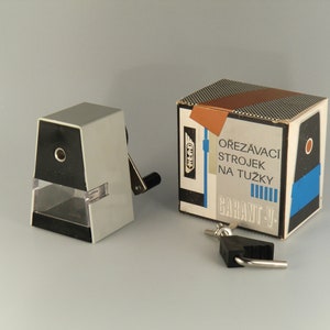 May include: A vintage gray and black pencil sharpener with a crank handle. The sharpener is in its original box, which is labeled "Ořezávací Strojek Na Tužky Garant V."