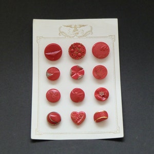 Puede incluir: Una tarjeta con doce botones rojos con detalles dorados. Los botones tienen diferentes diseños, incluyendo formas florales, geométricas y de corazón.