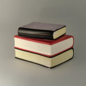 Op de afbeelding: Een stapel van drie keramische boeken. Het onderste boek heeft een gele paginarand, witte pagina's en een rode kaft. Het middelste boek heeft een zwarte paginarand, witte pagina's en een rode kaft. Het bovenste boek heeft een zwarte paginarand en een paarse kaft.