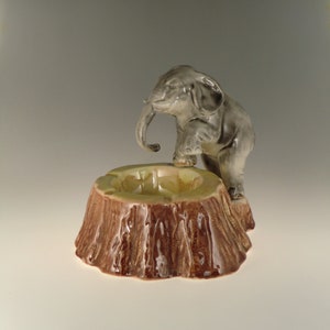 Peut inclure: Un cendrier en céramique en forme de souche d'arbre avec une petite figurine d'éléphant debout dessus. L'éléphant est gris et a sa trompe levée. Le cendrier est brun et a un glaçage vert.