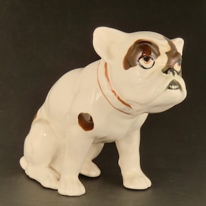 Può includere: Figurina in ceramica bianca di un bulldog francese con macchie marroni. Il cane è seduto con la testa girata di lato e le orecchie dritte.