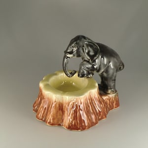 Puede incluir: Un cenicero de cerámica con forma de tronco de árbol con una pequeña figura de elefante de pie encima. El elefante es negro y gris y tiene la trompa en el cenicero. El cenicero es marrón y amarillo.