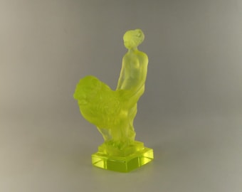 May include: A yellow glass figurine of a woman holding a basket. The figurine is standing on a square base.