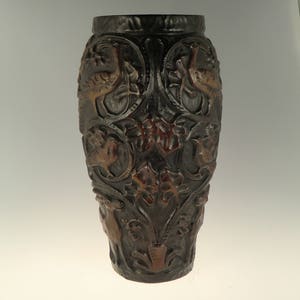 Puede incluir: Un jarrón de cerámica marrón oscuro con intrincados tallados de pájaros y diseños florales. El jarrón tiene una superficie texturizada y una forma ligeramente redondeada.