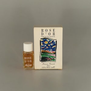 May include: A miniature bottle of Rose D'Or perfume and its accompanying box. The bottle has a white cap, and the box displays a colourful landscape painting. The text "Rose D'Or" is printed on both.