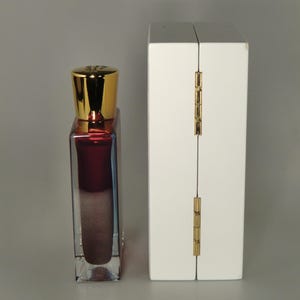 Op de afbeelding: Een klein, doorzichtig glazen flesje met een gouden dop en een donkerrode vloeistof erin. Het flesje staat naast een witte doos met een gouden scharnier.