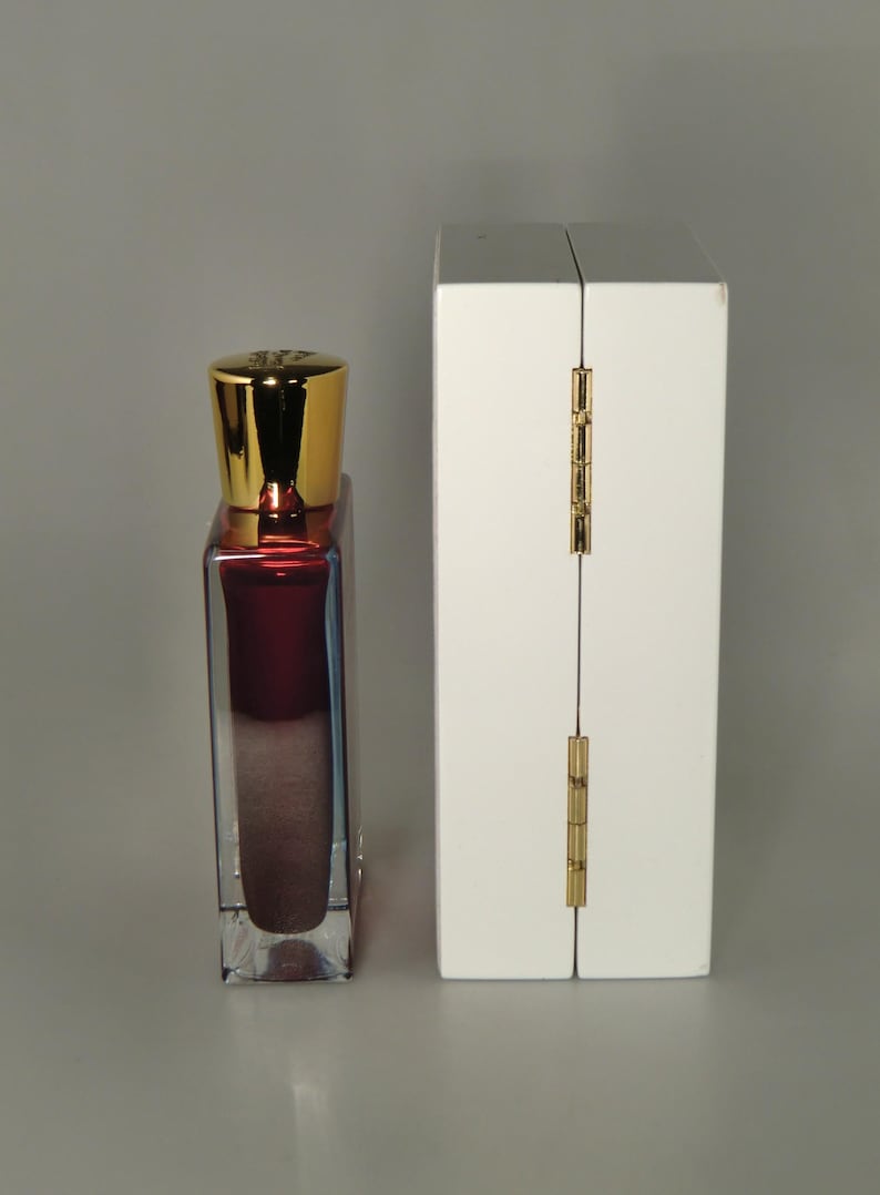 Op de afbeelding: Een klein, doorzichtig glazen flesje met een gouden dop en een donkerrode vloeistof erin. Het flesje staat naast een witte doos met een gouden scharnier.