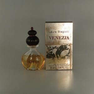 May include: A miniature bottle of Laura Biagiotti Venezia eau de parfum with a dark brown decorative cap and a matching box. The bottle contains a golden liquid. The box features the name "VENEZIA" in red lettering and the text "25 ml 0.84 FL. OZ."