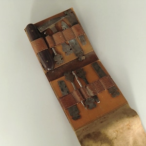 May include: An open, antique leather tool roll containing various metal tools. The roll is tan and brown, with tools secured by leather straps. A dark brown, cylindrical object is visible on the left side.