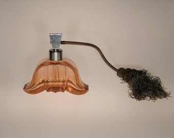 Può includere: Un flacone di profumo vintage con un corpo in vetro a forma di tulipano color pesca. Presenta una parte superiore in metallo argentato e un lungo stelo curvo che conduce a un'estremità scura e con nappe. La bottiglia ha delle scanalature verticali.
