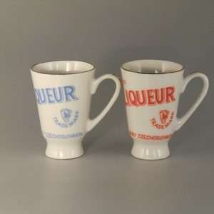 Könnte beinhalten: Zwei weiße Keramik-Likörbecher mit goldenen Rändern und Henkeln. Ein Becher hat blauen Text mit der Aufschrift "LIQUEUR", einem Wappen und "CZECHOSLOVAKIA". Der andere Becher hat roten Text mit dem gleichen Design.
