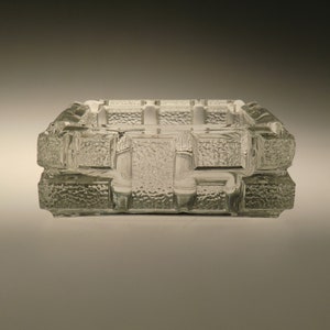 Puede incluir: Un cenicero de vidrio transparente con un diseño geométrico texturizado. El cenicero tiene forma cuadrada y un borde elevado.