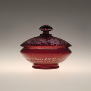 Könnte beinhalten: Eine rote Glasdose mit Deckel, verziert mit weißen Blumenmustern. Die Dose hat einen runden Körper und einen leicht ausgestellten Rand.