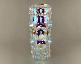 May include: Iridescent glass vase with a cylindrical shape. The vase features a textured surface with repeating patterns of rounded and bow-shaped designs. The glass reflects a spectrum of colors, including blue, purple, and yellow, creating a shimmering effect.
