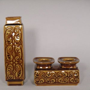 Może przedstawiać: Zestaw dwóch brązowych świeczników ceramicznych z ozdobnym wzorem kwiatowym. Świeczniki są prostokątne i mają dwa okrągłe pojemniki na świece. Świeczniki są wykonane z brązowej glazurowanej ceramiki.