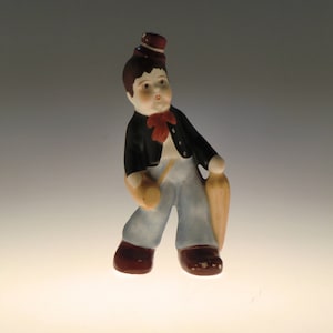 Peut inclure: Une figurine en porcelaine d'un garçon portant une veste noire, un pantalon bleu et un chapeau marron. Il tient une canne dans sa main droite et un parapluie dans sa main gauche.