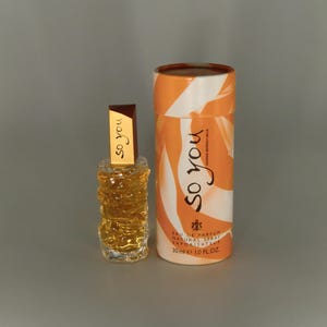 May include: A perfume bottle and its cylindrical box. The bottle is small, clear glass with a textured design and a brown cap. The box is white with orange swirls and the words "So You" in a script font.