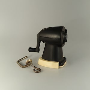 Peut inclure: Un taille-crayon noir avec une base blanche et une manivelle en métal. Le taille-crayon a un corps rond et un petit trou pour insérer des crayons.