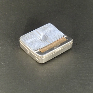 May include: A silver cigarette rolling machine with a textured surface and the word "RAPID" printed on the side.