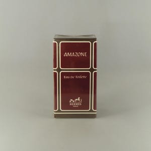 May include: A maroon-coloured box of "Amazone" Eau de Toilette. The box has a white border and text, with the brand name "Hermes Paris" at the bottom. The box is on a grey background.